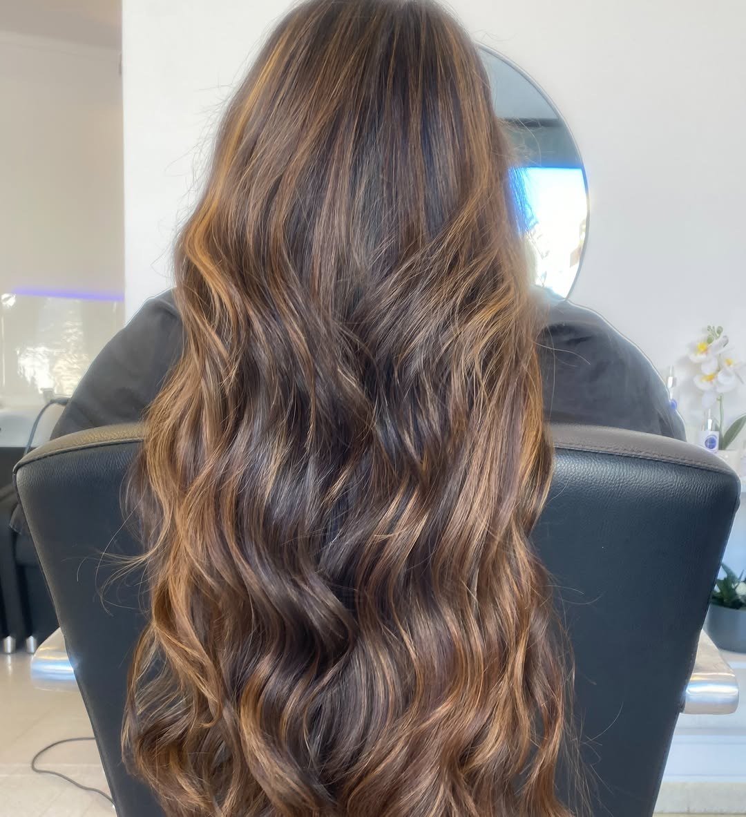Cabelo comprido castanho com balayage, visto de costas numa cadeira do salão, com um espelho redondo ao fundo