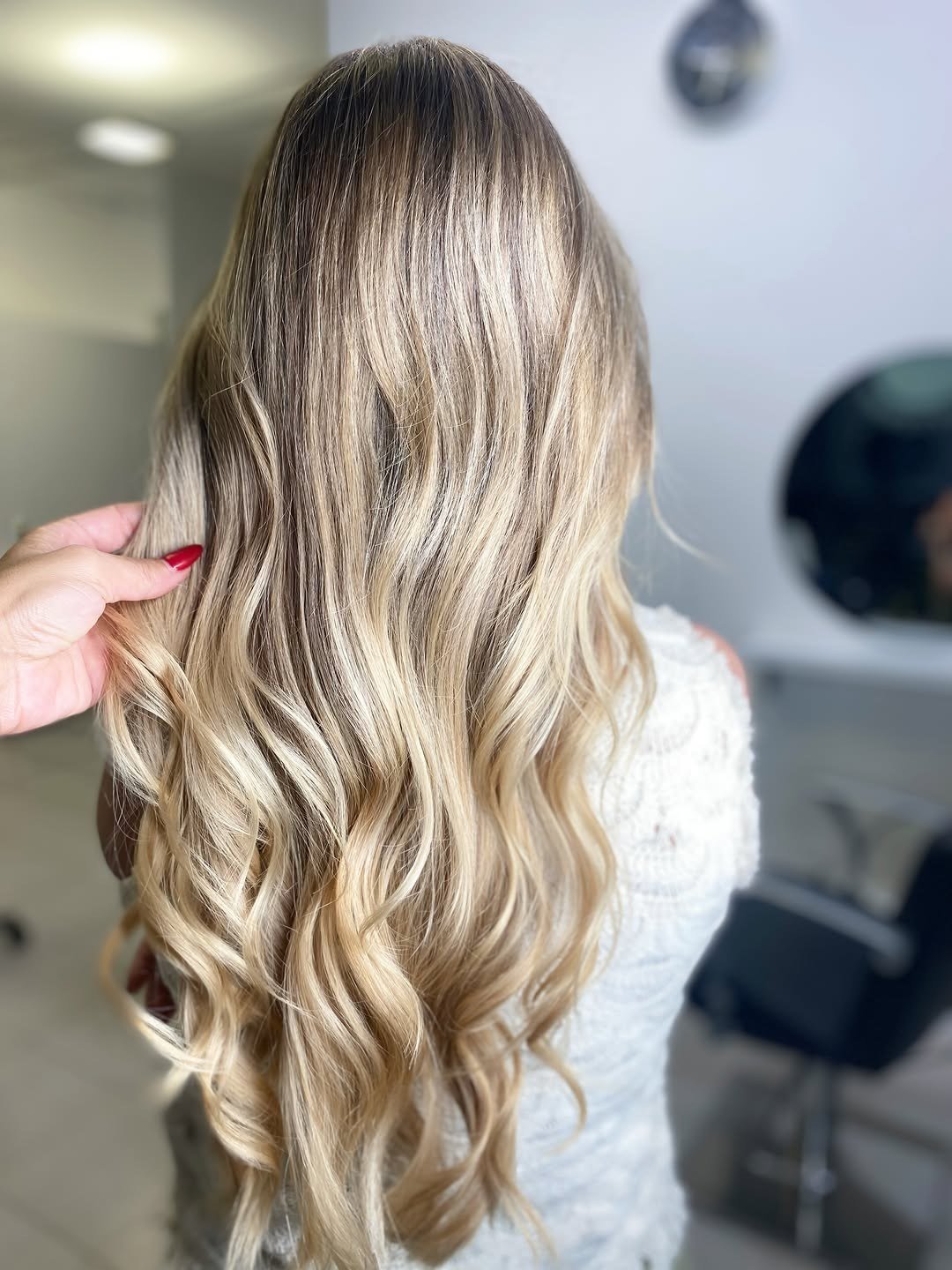 Cabelo comprido loiro claro com balayage e ondas, visto de costas, com uma mão a segurar uma madeixa